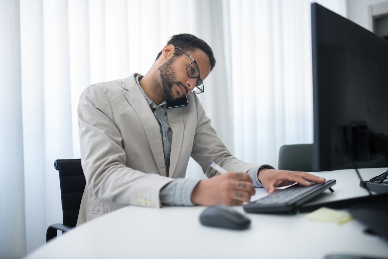 Focused businessman multitasking on phone and writing at office desk with computer.